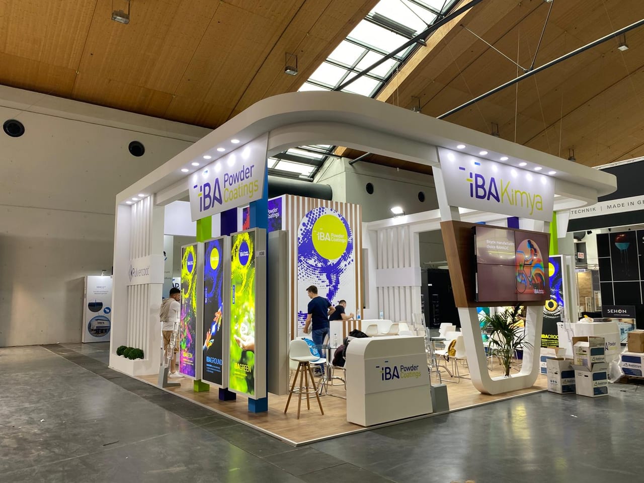İBA Kimya PaintExpo 2024 standının köşe görünümü; yuvarlatılmış beyaz alınlık, renkli aydınlatmalı ürün totemleri ve ahşap zemin.