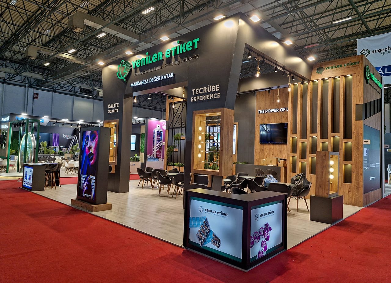 Aynı stand fuar salonunda kurulmuş hâlde; görselleştirmedeki alınlık, teşhir nişleri ve banko yerli yerinde duruyor.