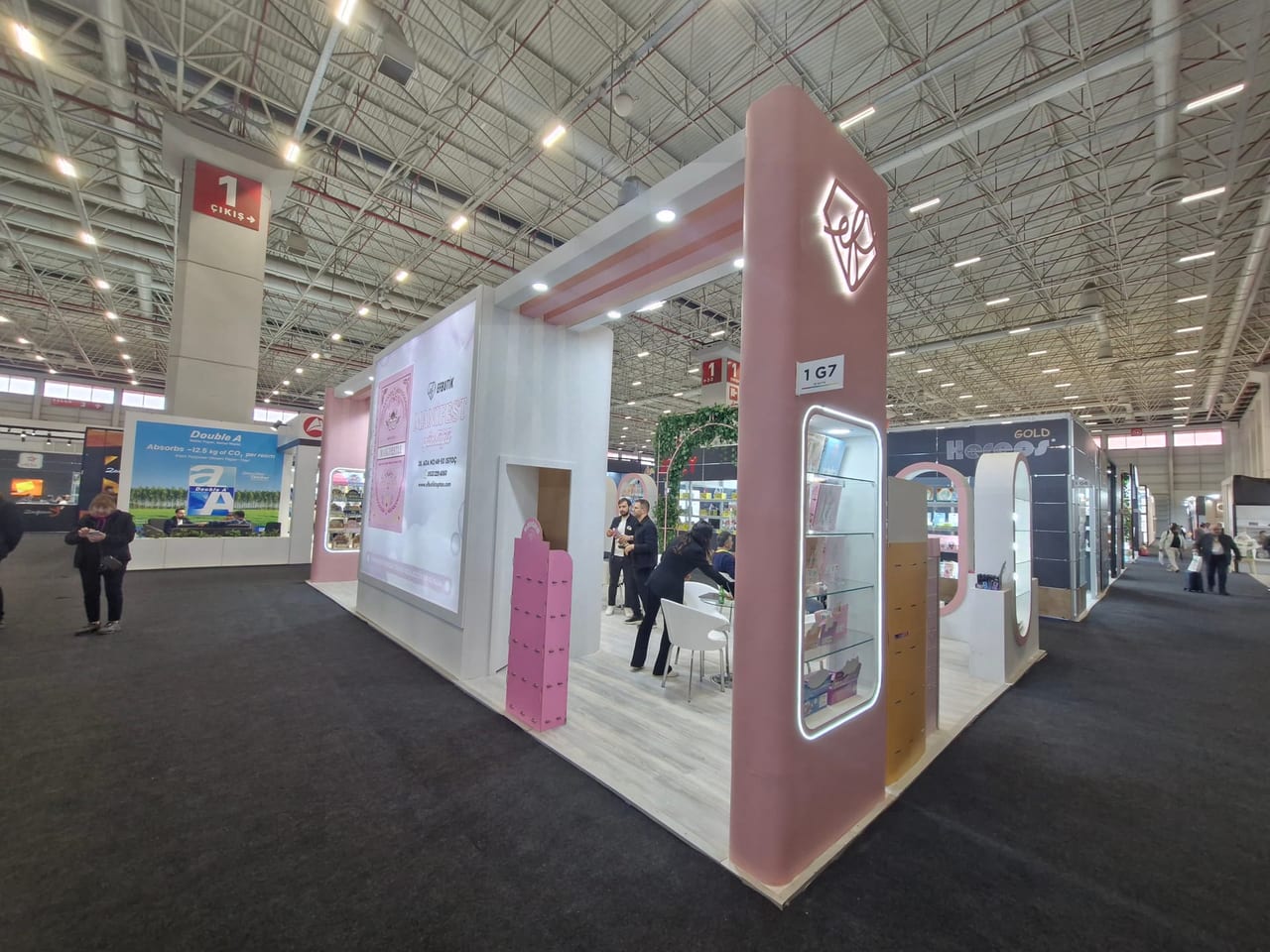 Fuar sürerken stand koridordan görünüyor; ziyaretçiler geziniyor ve görüşme alanı kullanılıyor.