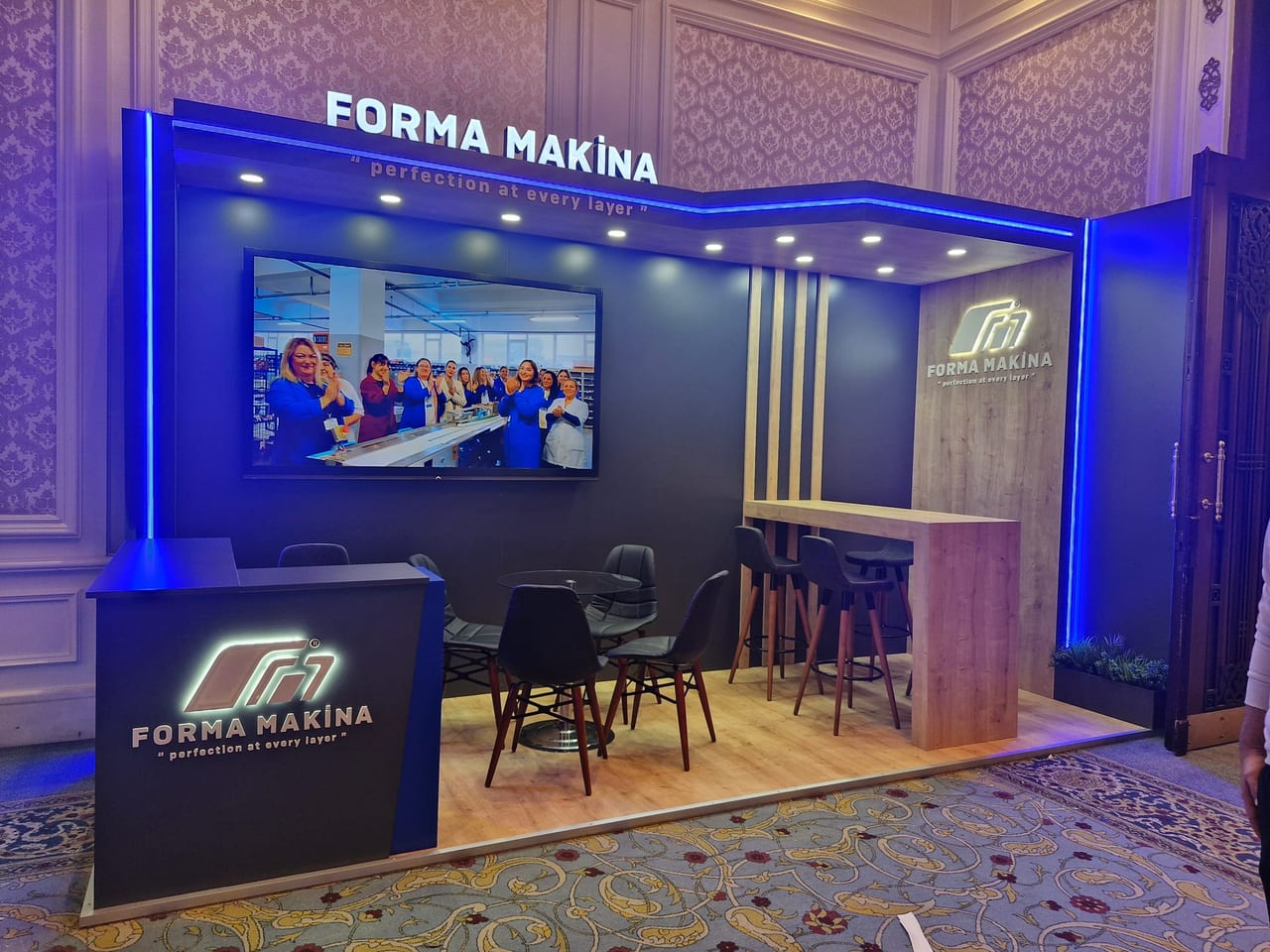 Forma Makina standı: aydınlatmalı alınlık ve banko logosu, ahşap kaplama panel, duvara gömülü ekran, önünde görüşme masası ve bar oturma grubu.