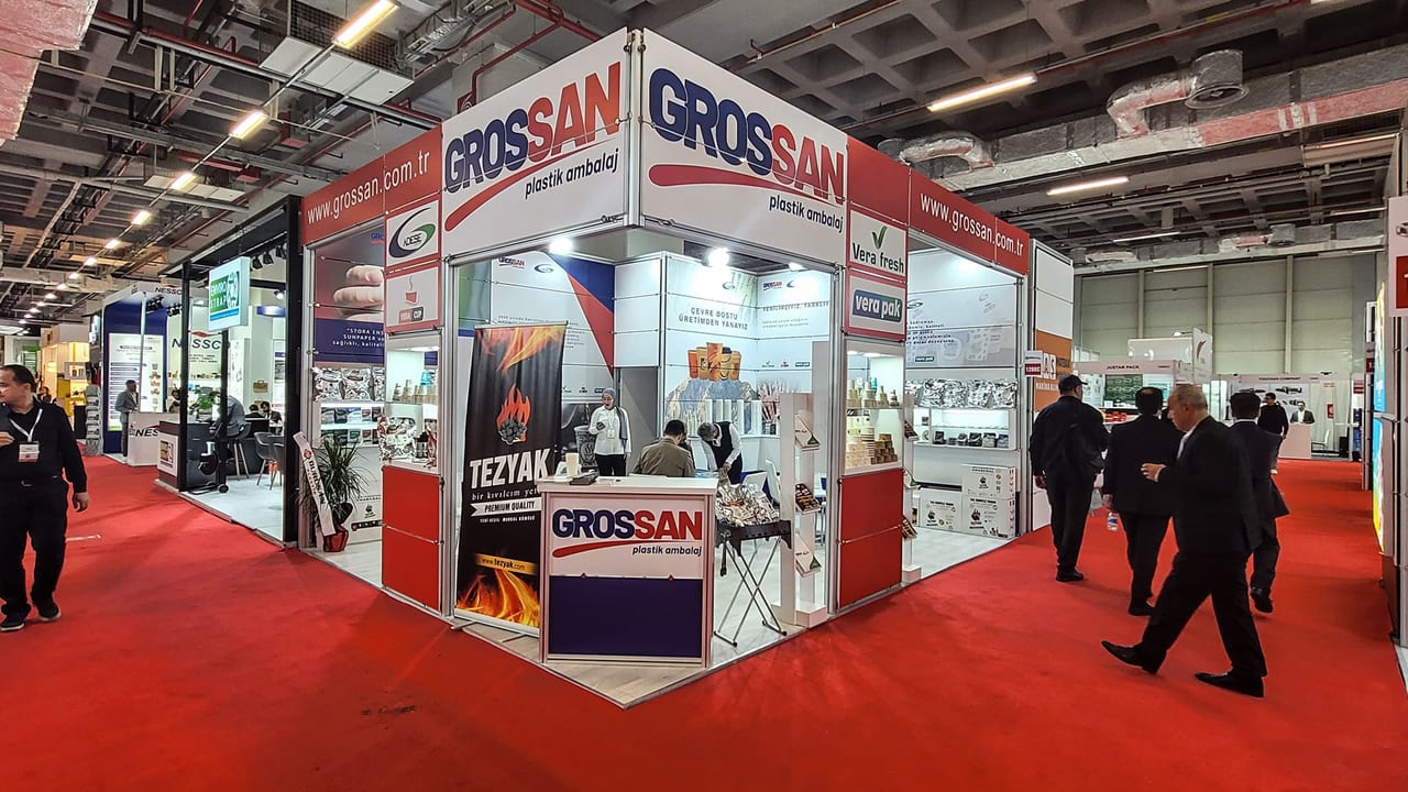 Grossan için kurduğumuz gıda ambalajı fuar standı: iki açık cepheli köşe stand, kırmızı-beyaz marka alınlıkları, ürün rafları ve karşılama bankosu.