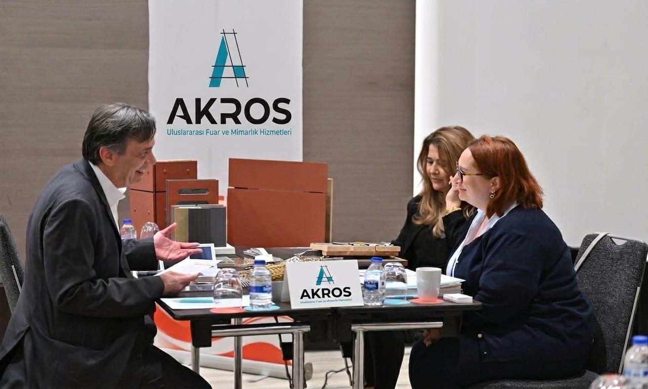 Akros ekibinden bir mimar iki katılımcıyla masa başında görüşüyor; masada ahşap ve kaplama malzeme örnekleri duruyor, arkada Akros afişi yer alıyor.