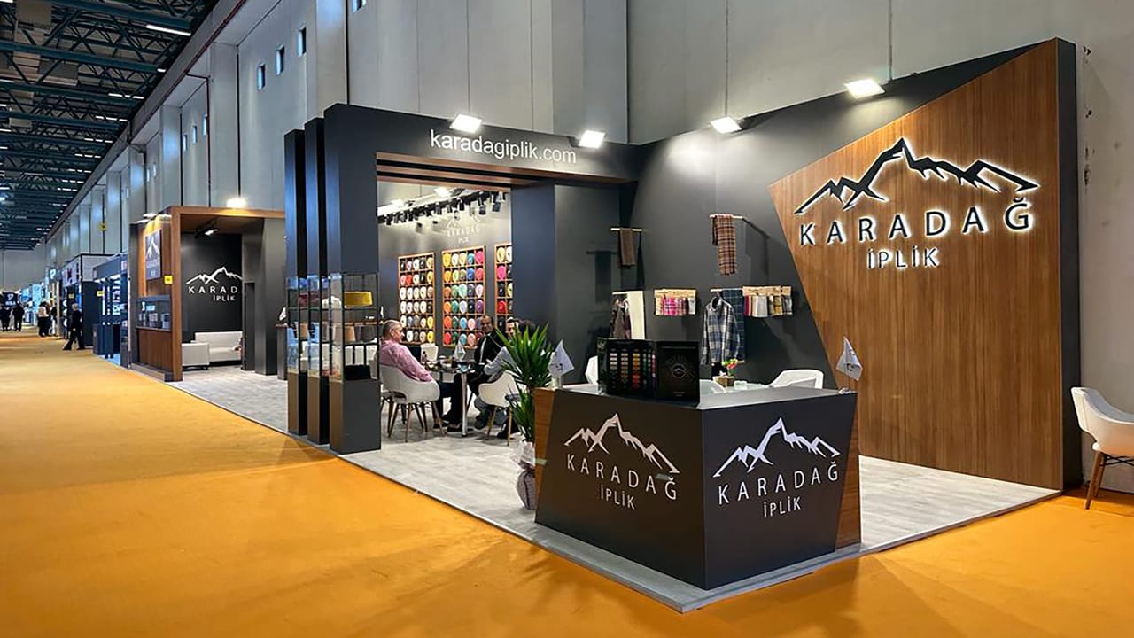 Karadağ İplik için kurduğumuz tekstil fuarı standı: ahşap kaplama marka duvarı üzerinde ışıklı logo, iplik makarası teşhir duvarı, cam vitrinler ve toplantı masası.